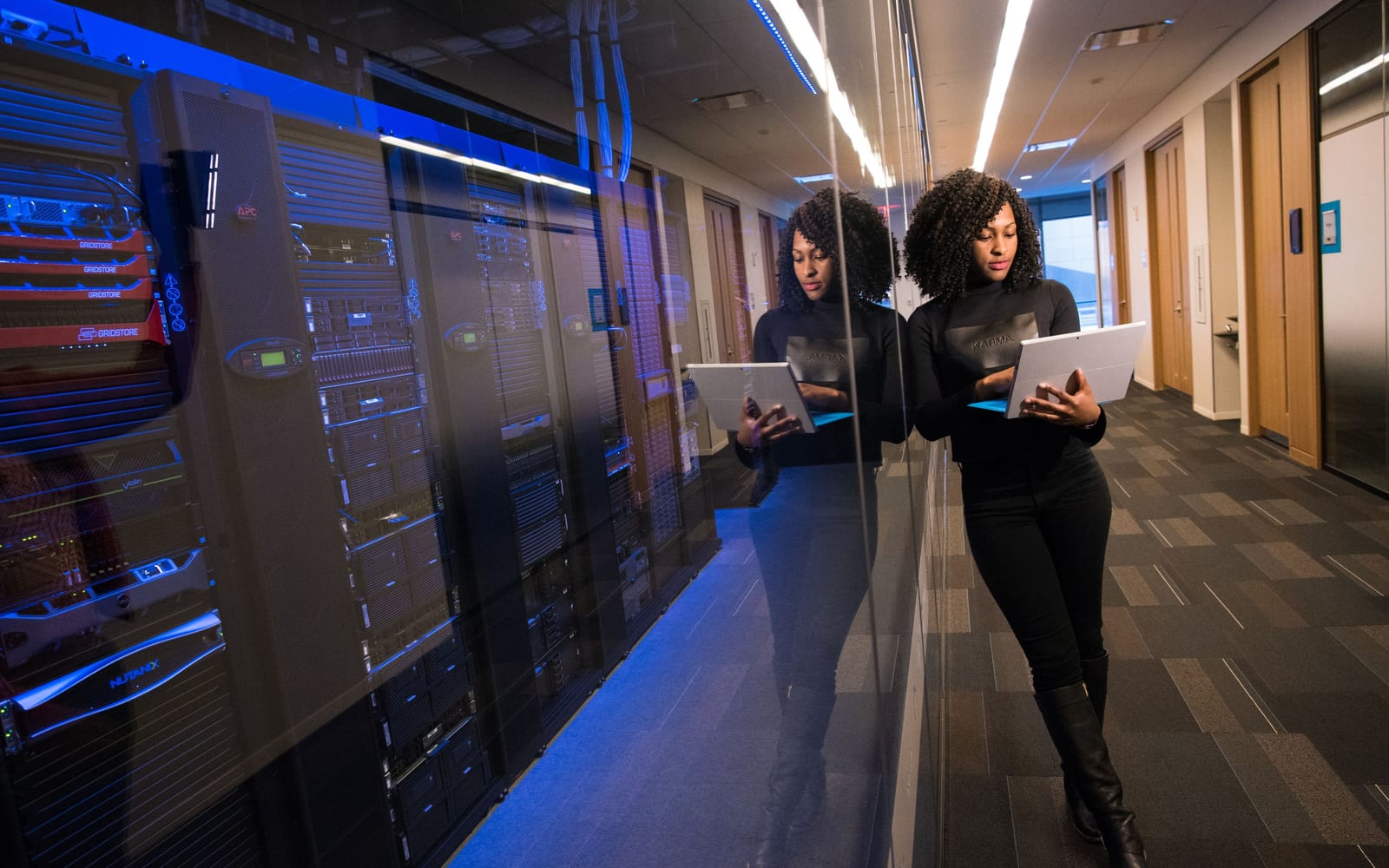 An engineer reading a laptop in an office corridor, a server room lit blue behind the glass wall beside her
